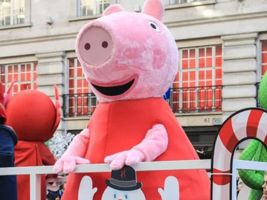 Peppa Pig float at a parade, surrounded by festive decorations and buildings in the background.