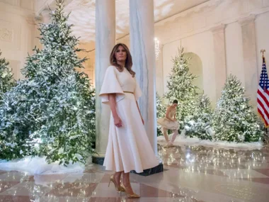 Elegant woman in white dress stands among sparkling Christmas trees in a decorated hall with an American flag visible.