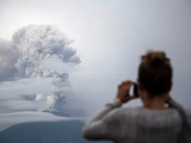 Person photographing erupting volcano with clouds of ash in the sky.