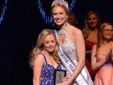 Two women at a beauty pageant, one holding a plaque, both smiling with others in the background.