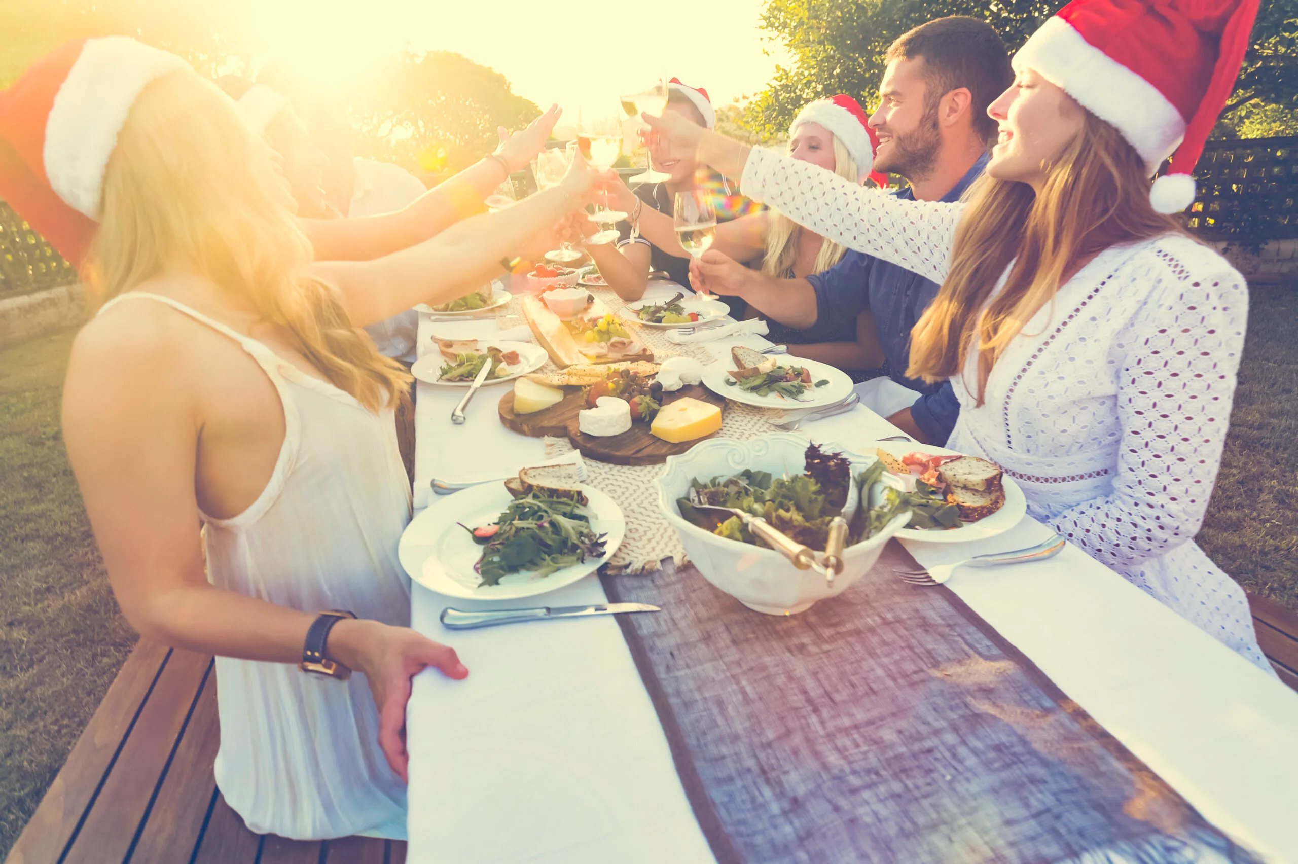 Group of people in Santa hats toasting at an outdoor holiday dinner party, with salads and cheese on the table.