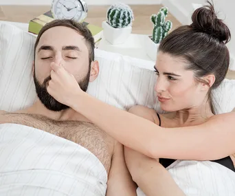 "Person in bed pinching another's nose, cactus plants and clock in background."