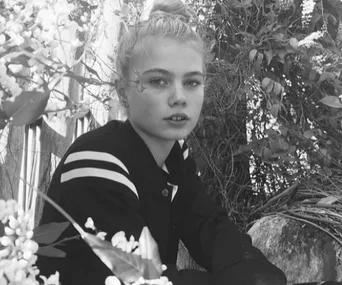Young girl seated in a garden setting, surrounded by foliage and flowers, looking towards the camera. Black and white photo.