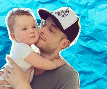 Man in a cap gently holding a baby against a crumpled blue background.