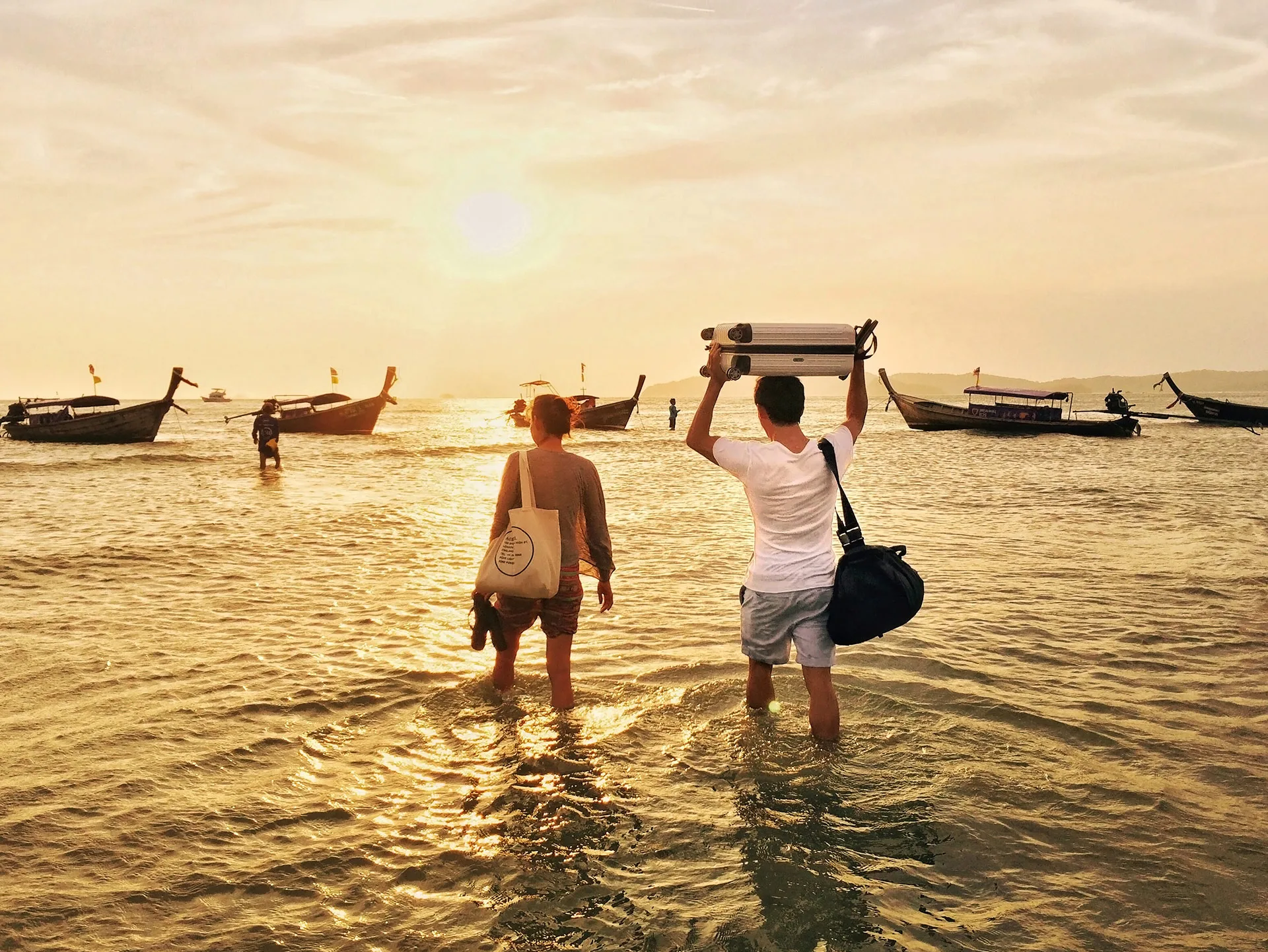 Two people walking through shallow water towards boats at sunset, carrying bags and a suitcase overhead.