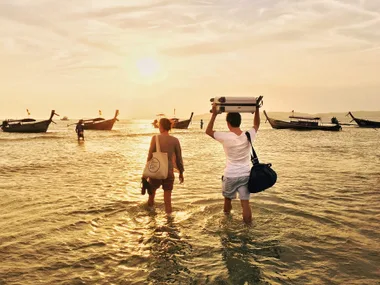 Two people walking through shallow water towards boats at sunset, carrying bags and a suitcase overhead.