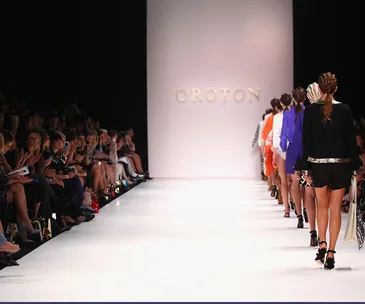 Models walking on a runway with "Oroton" displayed in the background, audience seated on both sides.