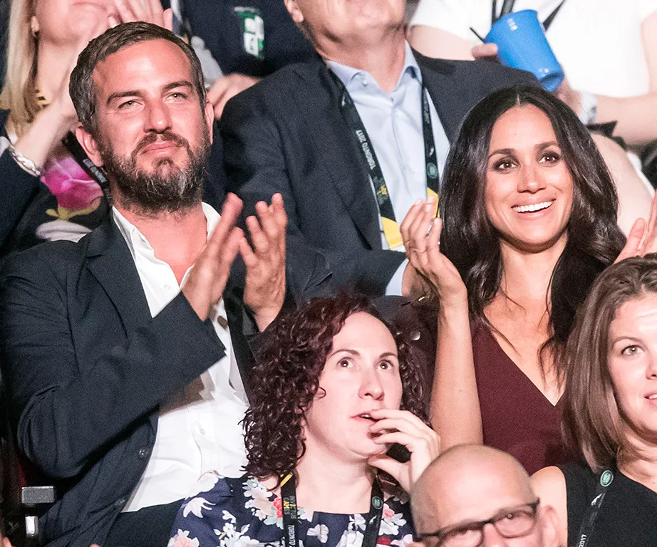 A smiling group at an event, with a man in a suit clapping and a woman in a dark top seated beside him.