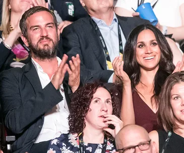 A smiling group at an event, with a man in a suit clapping and a woman in a dark top seated beside him.