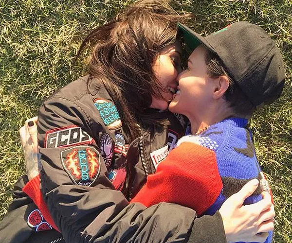 Two individuals lying on grass, smiling, wearing colorful jackets, embracing closely.