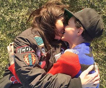 Two individuals lying on grass, smiling, wearing colorful jackets, embracing closely.