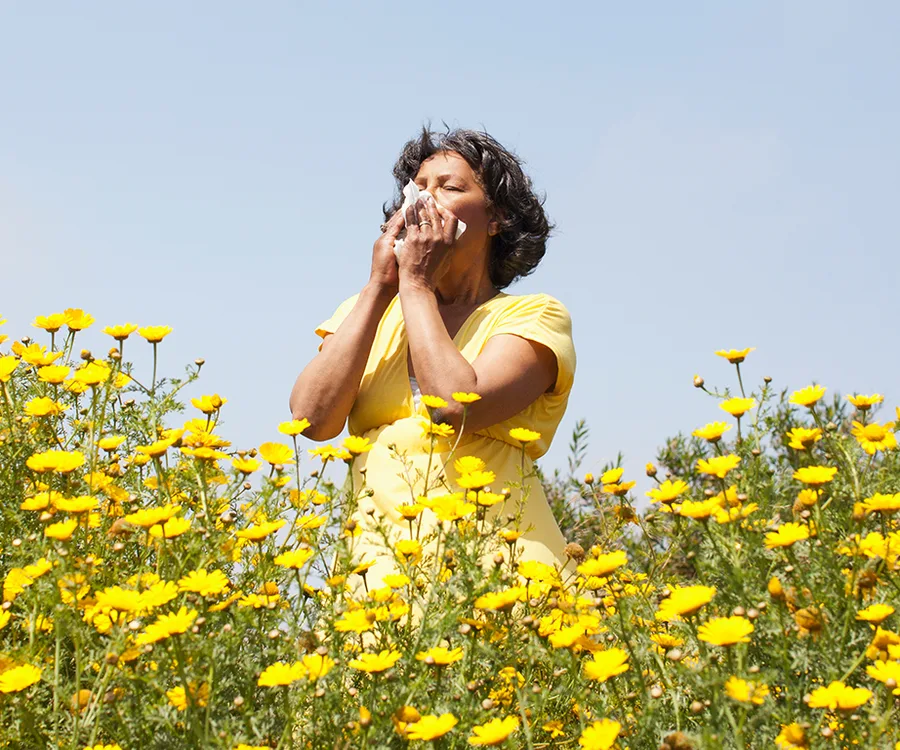 A woman in a yellow shirt sneezing into a tissue in a field of yellow flowers under a clear blue sky.