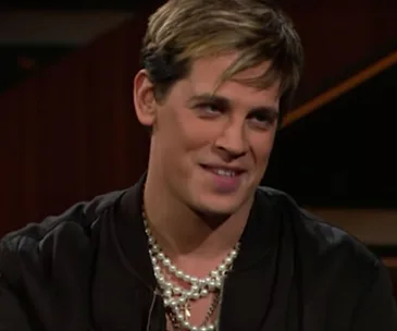 A person with short, light brown hair, wearing a black jacket and multiple pearl necklaces, smiling slightly.