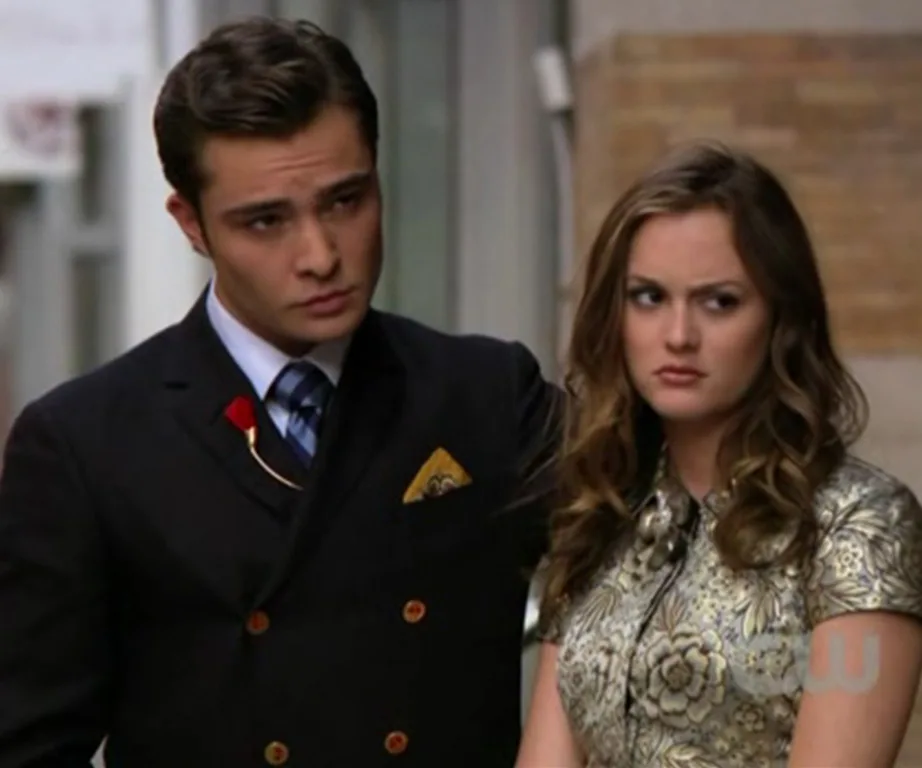 A well-dressed couple stands outdoors; the woman looks displeased.