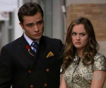 A well-dressed couple stands outdoors; the woman looks displeased.