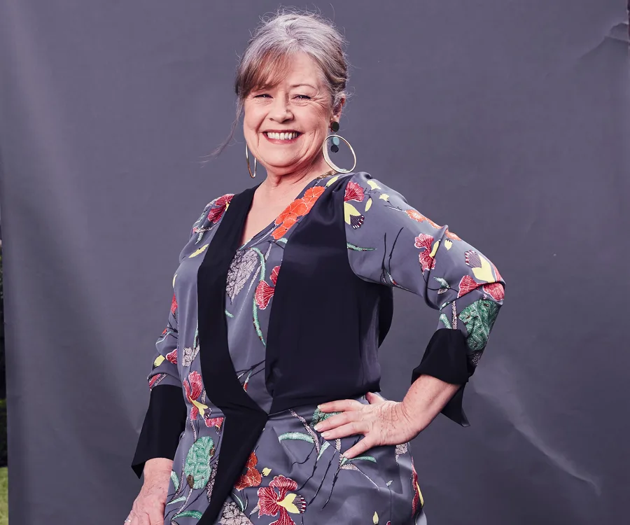 A woman smiling, wearing a floral-patterned blouse with hoop earrings, standing against a gray background.