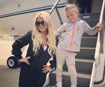 "Jessica Simpson and daughter posing on airplane stairs, both smiling, with the daughter in a floral tracksuit."