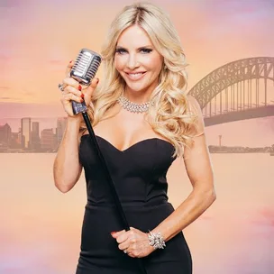 Blonde woman in a black dress holding a microphone against a sunset city skyline with a bridge in the background.