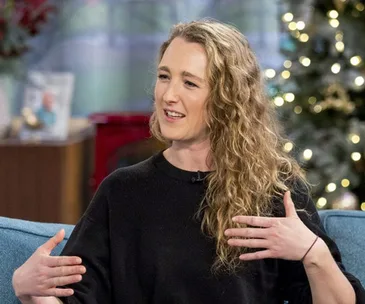 A woman with curly hair in casual clothes gestures while speaking on a talk show set decorated for the holidays.