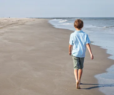 A young boy walks alone on a sandy beach, wearing a blue shirt and shorts, with the ocean to his right.