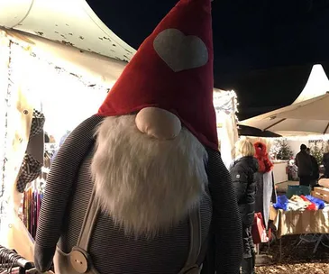 Gnome statue with red hat and heart at a Christmas market stall, surrounded by festive lights and people.