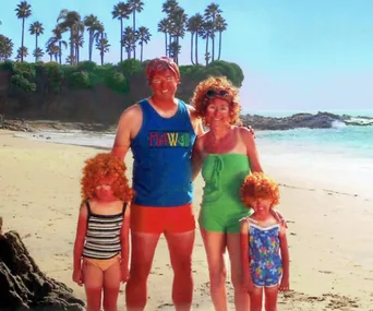 Family in swimsuits with orange wigs posing on a sunny beach, palm trees in the background.