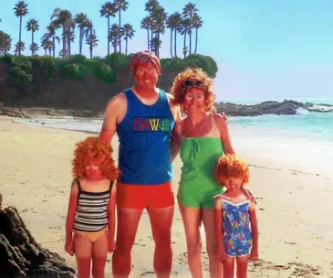 Family in swimsuits with orange wigs posing on a sunny beach, palm trees in the background.