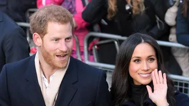 Couple smiling and waving at a public event, surrounded by a crowd and fences.