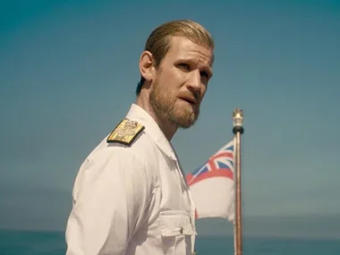 Officer in uniform, standing on a boat with a British flag, under clear blue sky.