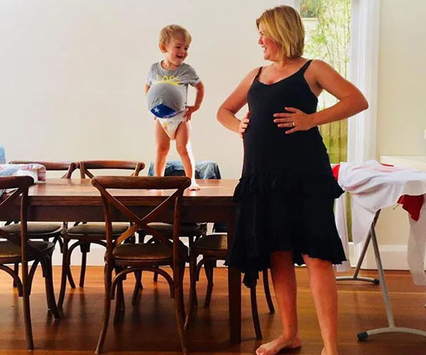 Pregnant woman smiles at a child standing on a table, both mimicking pregnant bellies in a bright room.