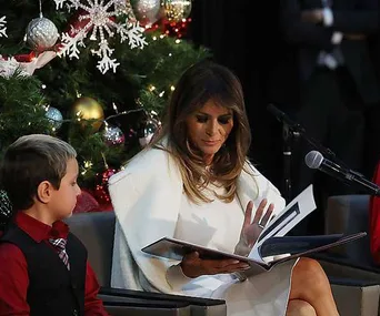 Melania Trump reads a book to children by a decorated Christmas tree.