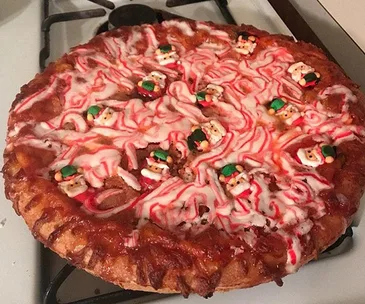 A pizza with red and white icing, topped with small Santa decorations, on a stove, resembling a Christmas baking fail.