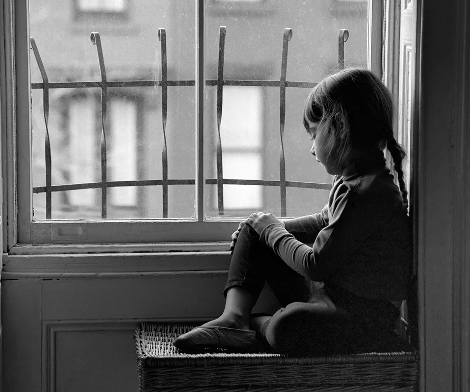 Child sitting by a window, looking outside with a contemplative expression; black and white photo.