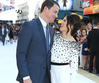 A man in a suit and a woman in a white dress smiling at each other on a red carpet.