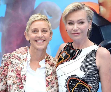 Two individuals smiling at an event, one wearing a floral blazer and the other in a patterned dress, with a colorful backdrop.
