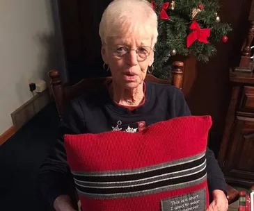 An elderly woman holds a red pillow made from a shirt, sitting by a Christmas tree.
