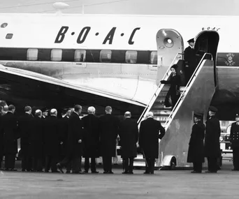 A woman descends stairs from a BOAC airplane, greeted by a line of formally dressed men on the tarmac.