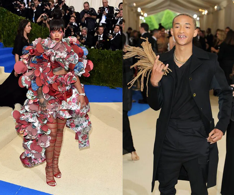 Two individuals at a fashion event: one in a floral avant-garde dress, the other in a black suit holding hair and wearing grills.