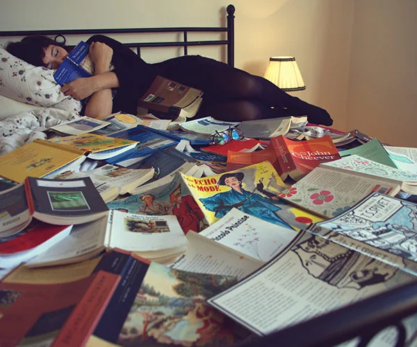 Woman sleeping on a bed covered with open books, holding one in her hand.
