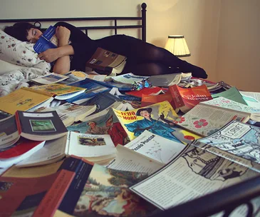 Woman sleeping on a bed covered with open books, holding one in her hand.