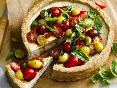 Gluten-free savory tart topped with colorful cherry tomatoes and basil leaves, one slice cut out on parchment paper.