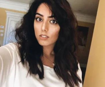 A woman with long dark hair and a white top takes a selfie indoors, looking directly at the camera.