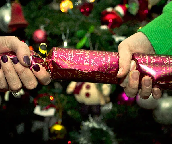 Two hands pulling a Christmas cracker in front of a decorated Christmas tree.