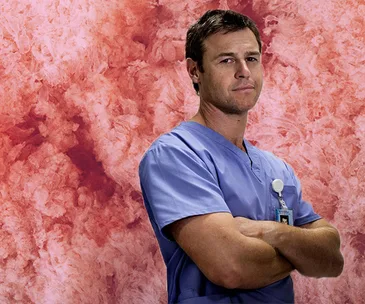 Surgeon in blue scrubs stands with arms crossed against a textured red background.