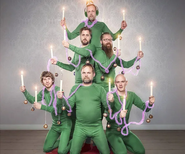 A group of six men dressed in green form a human Christmas tree, holding candles and purple tinsel.