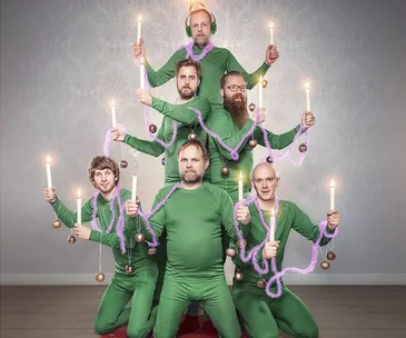 A group of six men dressed in green form a human Christmas tree, holding candles and purple tinsel.