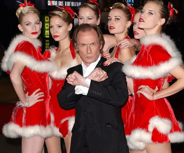 Bill Nighy poses with women in red Santa dresses, recreating a scene from "Love Actually."