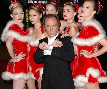 Bill Nighy poses with women in red Santa dresses, recreating a scene from "Love Actually."