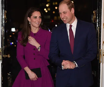 Prince William and Duchess Kate attending a formal event, she wears a purple dress, he wears a blue suit with red tie.