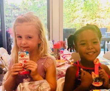 Two young girls smiling, one holding a juice box, the other holding a spoon, both at a colorful children's party.